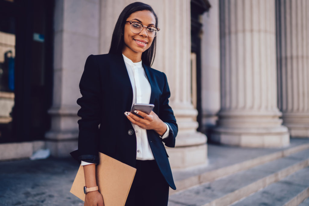 Portrait of successful African American businesswoman dressed in stylish suit holding in hand folder and mobile phone while standing outdoors near financial office, young woman lawyer using smartphone