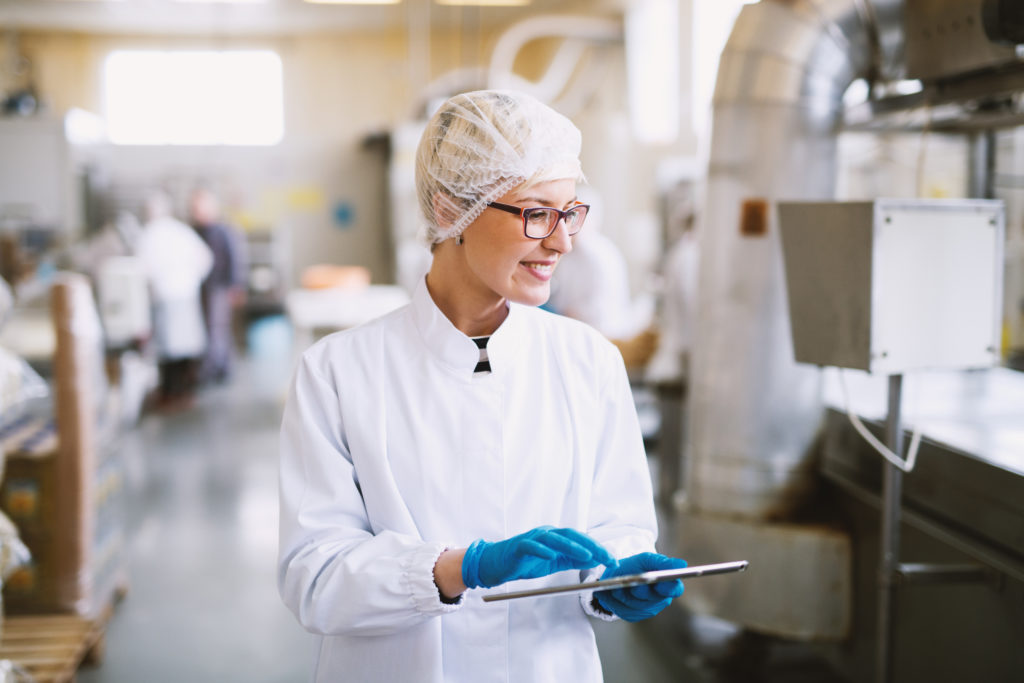 Smiley female worker in sterile clothes using tablet and checking how production line is working.