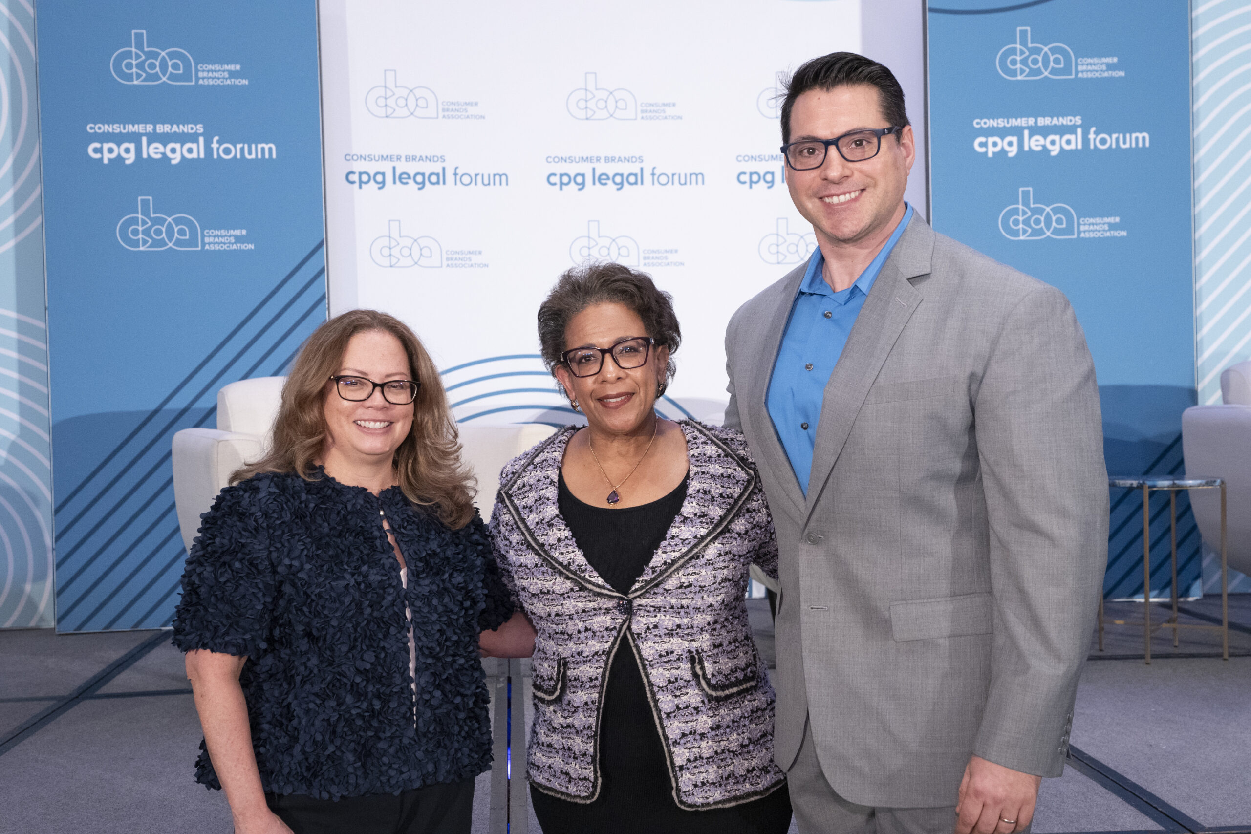 Consumer Brands COO and General Counsel Stacy Papadopoulos (L) and Consumer Brands VP and Deputy General Counsel Joseph Aquilina (R) stand on either side of former U.S. Attorney General Loretta Lynch to pose for a photo during the CPG Legal Forum. They are standing in front of a blue and white paneled background with the Consumer Brands logo on it.